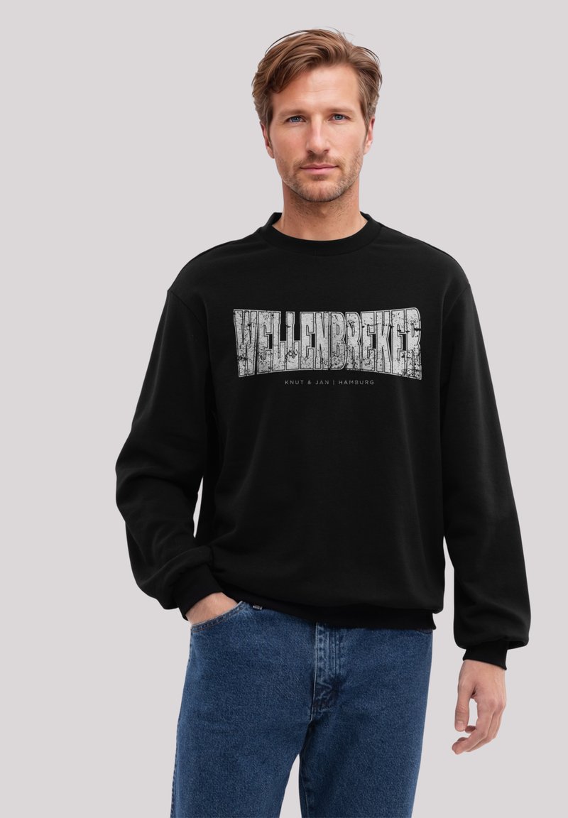Schwarzer Sweatshirt mit Rundhalsausschnitt, versehen mit einem großen weißen grafischen Text "WELLENBREKER" und kleinerem Text darunter. Kombiniert mit blauen Jeans.