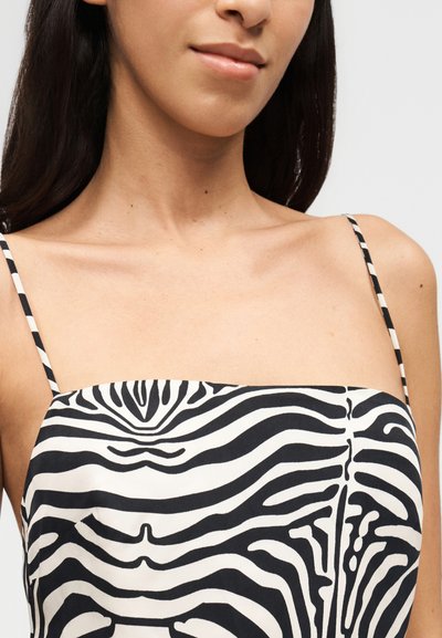 Γυναίκα που φοράει μαύρη και λευκή μπλούζα χωρίς μανίκια με ρίγες ζέβρας και λεπτές τιράντες, εμφανίζοντας το λαιμό, τους ώμους και ένα διακριτικό χαμόγελο.