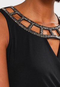 Frau trägt ein ärmelloses schwarzes Oberteil mit einem silbernen, mit Perlen besetzten geometrischen Ausschnitt.