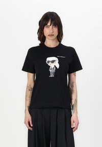 Juodas medvilninis marškinėliai su animacine grafika, vaizduojančia figūrą su akinių nuo saulės ir kostiumu, virš iliustracijos užrašyta "KARL LAGERFELD".
