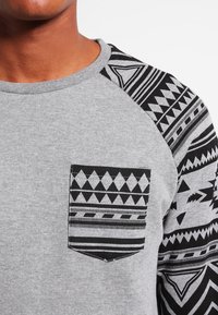 Sudadera gris de algodón con manga negra con estampado. Cuenta con un bolsillo en el pecho izquierdo con diseños geométricos, texturas contrastantes y detalles de costura.