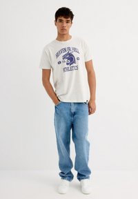 Camiseta de algodón gris con el estampado "HEAVEN OR HELL ATHLETICS" y gráficos en azul, combinada con jeans de mezclilla azul claro y zapatillas blancas.