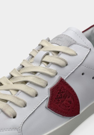 Λευκό δερμάτινο αθλητικό παπούτσι με εκρού κορδόνια, που διαθέτει ένα υφασμάτινο κόκκινο έμβλημα στο πλάι και μια λεία γκρι γλώσσα.