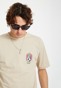 Beige short-sleeve t-shirt featuring a colorful logo with flame accents on the chest. Accessories include black sunglasses and a silver necklace.