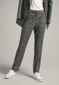 Personne portant un pantalon de costume à carreaux gris et une veste assortie avec des baskets blanches, se tenant devant un fond gris clair uni.