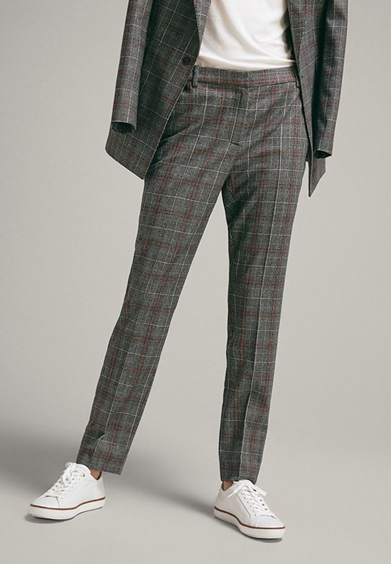 Personne portant un pantalon de costume à carreaux gris et une veste assortie avec des baskets blanches, se tenant devant un fond gris clair uni.