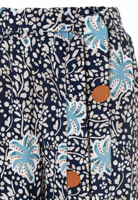 Maritimer, floralbedruckter Stoff in Dunkelblau mit hellbeigen Blättern und leuchtend blauen Palmmotiven; verfügt über Falten und ein braunes, rundes Akzentdetail in der Nähe des Taillenbundes.