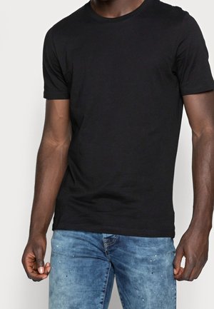 Man wearing a plain black short-sleeve t-shirt and blue denim jeans with paint splatter details, standing against a white background.