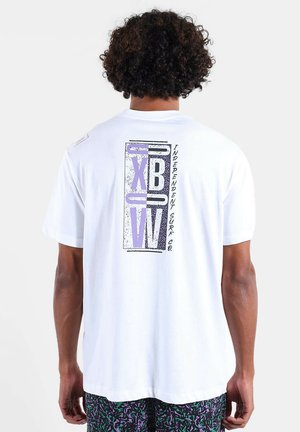 Homme aux cheveux bouclés portant un t-shirt blanc avec un texte graphique violet et noir "XB W" et "Independent Surf Co." dans le dos.