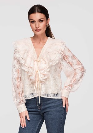 Femme portant un chemisier en dentelle crème avec un col volanté et un détail de nouage, assorti à un jean bleu foncé, posant devant un fond neutre.