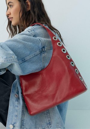 Femeie cu păr lung și închis la culoare, purtând o geacă ușoară din denim și având pe umăr o geantă roșie de piele cu detalii argintii tip ochiuri.