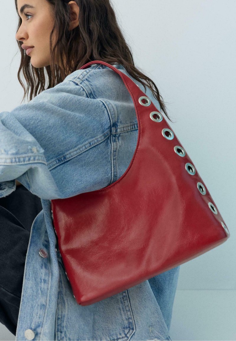 Femme aux longs cheveux bruns portant une veste en jean claire, tenant un sac à main en cuir rouge avec des détails en œillets argentés sur son épaule.