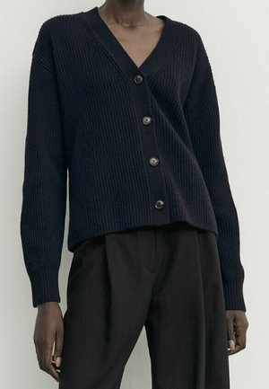 Cardigan noir côtelé à quatre boutons, porté sur un pantalon noir taille haute, sur une personne debout devant un fond uni.