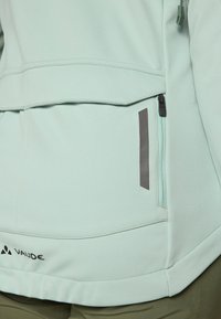 Nærbilde av en lys mintgrønn jakke med sidelomme med glidelås og grå detaljer, med en synlig merkevarelogo ved kanten.