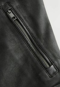Tissu en cuir noir présentant une surface texturée avec une fermeture éclair en métal argenté sur le côté, mettant en valeur un design angulaire et des détails cousus.