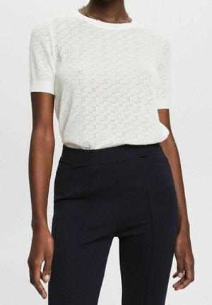 Dark-skinned person wearing a white short-sleeve knit top with eyelet pattern and high-waisted black pants, with a silver chain necklace.