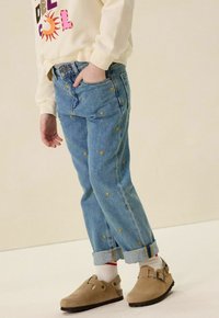 Jeans en denim bleu clair avec broderie en forme de cœur jaune, manches retroussées et poches latérales. Associés à des chaussures beiges et des chaussettes blanches avec des rayures rouges.