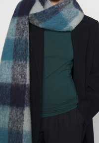 Blå och grå rutig halsduk av mjukt, strukturert tyg hängande över en mörk kappa, med en underliggande mörkgrön topp synlig.
