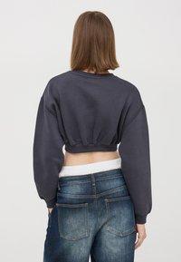 Skrócona, węglowa bluza z długimi, bufiastej rękami i elastycznym wykończeniem. Noszona z niebieskimi dżinsami o wyblakłym wykończeniu i tylnych kieszeniach.