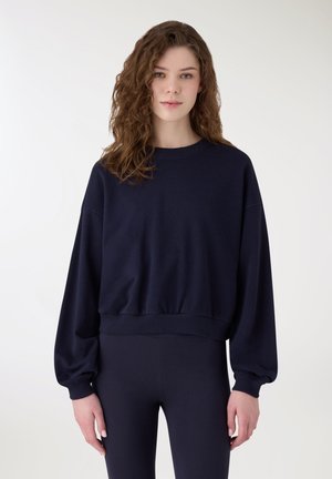 Giovane donna con capelli ricci castani che indossa una felpa blu navy larga e leggings aderenti blu navy, in piedi davanti a uno sfondo bianco semplice.