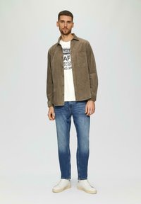 Bruine corduroy overhemden, wit graphic t-shirt, licht beschadigde blauwe jeans, en witte sneakers op een effen achtergrond.