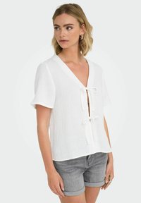 Vit blus med korta ärmar, V-hals och knytning framtill. Texturerad tyg med avslappnad passform, kombinerad med gråa shorts.