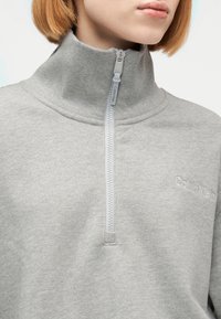 Κοντινό πλάνο ενός ατόμου που φοράει ανοιχτό γκρι φούτερ Calvin Klein με γιακά μισής ζώνης φερμουάρ κλειστό, εμφανίζοντας το λογότυπο της μάρκας στο στήθος.