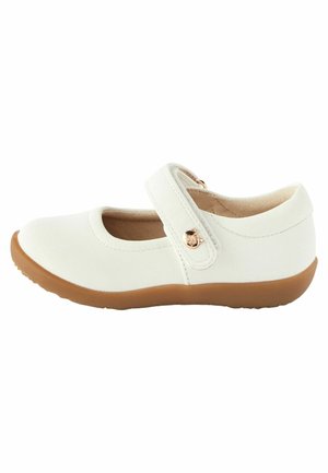 Chaussure Mary Jane blanche en cuir avec un bout arrondi, semelle plate en caoutchouc marron, bride ajustable et accent métallique circulaire.