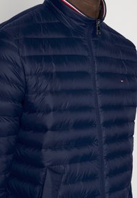 Granatowa kurtka puchowa z poziomym pikowaniem, wysokim kołnierzem i pełnym zamkiem błyskawicznym. Posiada małe logo na piersi.