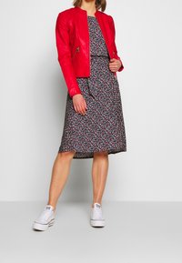 Veste bomber en cuir rouge sur une robe à motif floral avec une taille cintrée, assortie de baskets blanches. Texture lisse et couleurs vives.