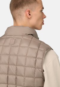 Beige pufferväst med ett kviltat mönster, ståkrage och logodetaljer. Materialet verkar glänsande och vadderat för isolering.