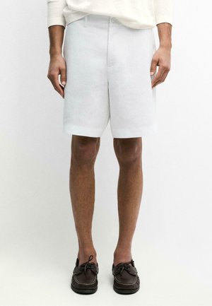 Mann trägt weiße, maßgeschneiderte Shorts und dunkelbraune Leder-Bootsschuhe, steht vor einem schlichten hellen Hintergrund.