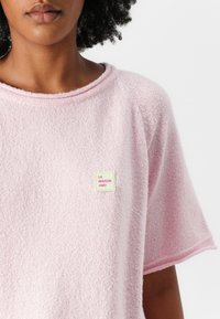 Rosa kortermet topp laget av teksturert stoff. Har en rund hals og en liten etikett med "LA MAISON AMV" på forsiden.