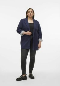 Marineblazer mit einem entspannten Schnitt und umgeschlagenen gestreiften Bündchen, kombiniert mit schwarzen Skinny-Jeans und schwarzen Slippern. Schlichte Hintergrund.