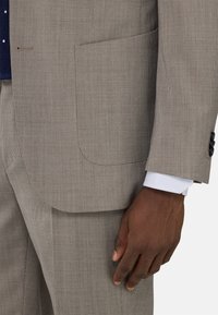 Tejido de traje a cuadros beige con superficie texturizada, que presenta un bolsillo en el pecho y puños abotonados. Puño de camisa a cuadros blanca visible.