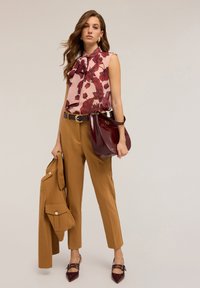 Donna che indossa un blusa senza maniche floreale rosa, pantaloni beige, tiene in mano una giacca beige abbinata e una borsa bordeaux, in piedi contro uno sfondo neutro.