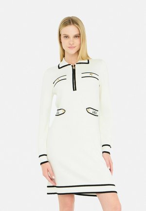 Abito bianco lavorato a maglia con bordi neri. Presenta un colletto con zip, due tasche frontali e dettagli a righe ai polsini e all'orlo. Texture liscia.