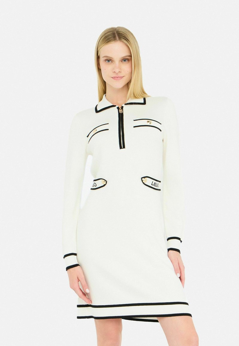 Abito bianco lavorato a maglia con bordi neri. Presenta un colletto con zip, due tasche frontali e dettagli a righe ai polsini e all'orlo. Texture liscia.