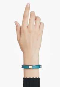 Genomskinligt turkoss armband med klara accenter, med fyra hexagonala utsmyckningar och en slät yta, bärs på en hand.