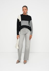 Chunky gebreide trui in zwart, wit en grijs kleurblokontwerp; gecombineerd met lichtgrijze wijde jeans en neutrale schoenen. Accessoires omvatten een kleine witte handtas.