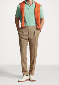 Light green polo shirt, orange cable-knit sweater draped, brown check-patterned trousers, and beige lace-up shoes. Clear lines and textures.