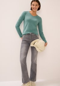 Langärmliges Teal-Oberteil, hellgraue Schlagjeans und eine crèmefarbene Tasche. Das Outfit zeichnet sich durch weiche Texturen und einen lässigen Schnitt aus.