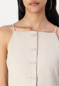 Camiseta sin mangas de color beige claro con tirantes finos, escote cuadrado y cinco botones a juego en la parte delantera. Tejido texturizado con un acabado suave.
