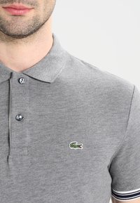Polo gris en coton texturé. Il présente une patte de boutonnage à deux boutons, un logo de crocodile vert et des accents rayés sur les manches.