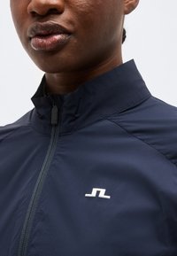 Κοντινό πλάνο ενός ατόμου που φοράει μπουφάν με φερμουάρ σε σκούρο μπλε χρώμα, με ψηλό γιακά και μικρό λευκό γεωμετρικό λογότυπο στο στήθος.