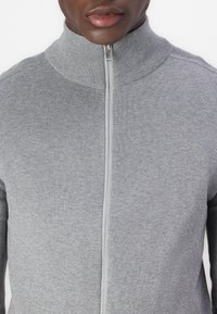 Sweater cinzento com fecho éclair, textura canelada, gola alta, detalhe de fecho de zíper prateado, design justificado, material suave, adequado para sobreposição em vários ambientes.