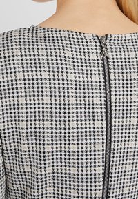 Vista trasera de una persona que lleva un vestido de cuadros en blanco y negro con un cinturón negro visible que corre por la parte superior de la espalda.
