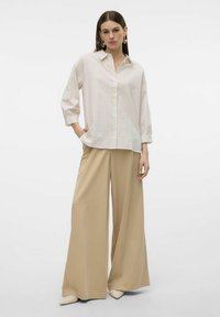 Camicia a righe bianche e rosa pallido con colletto e maniche lunghe, abbinata a pantaloni beige a gamba larga e scarpe a punta.
