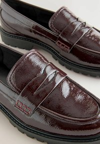 Mocassins en cuir verni bordeaux avec un design de couture surélevée, semelle en caoutchouc et détail de bande décorative sur le dessus.