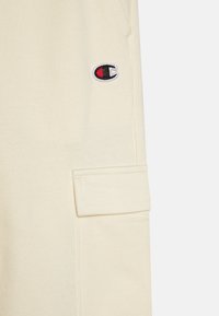 Kremfargede joggebukser med sidelomme og brodert logo med røde detaljer. Laget av myk tekstil med en jevn overflate.